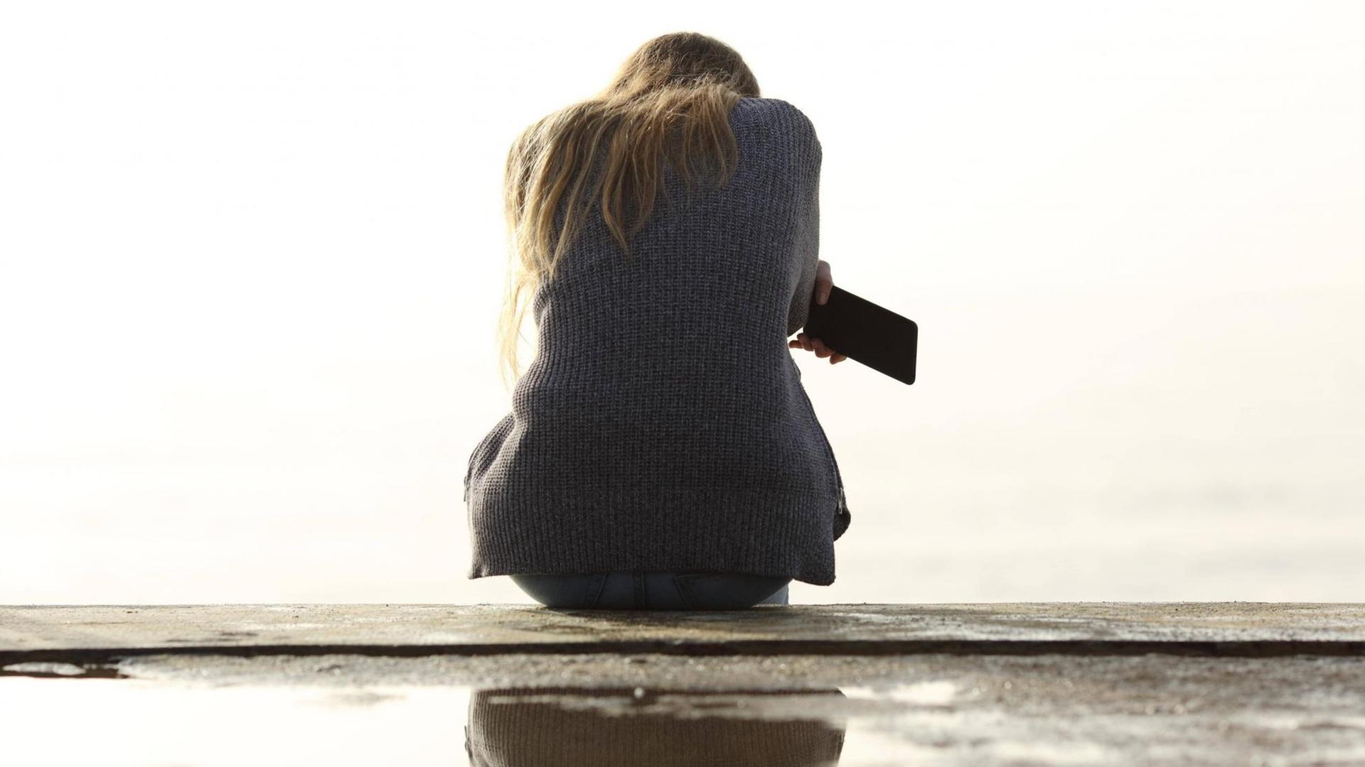 Ein Mädchen sitzt alleine. Ihr Rücken ist dem Betrachter zugewandt. Sie hält ein Smartphone in der Hand. Ein Mädchen sitzt alleine. Ihr Rücken ist dem Betrachter zugewandt. Sie hält ein Smartphone in der Hand.