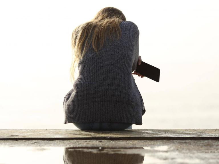 Ein Mädchen sitzt alleine. Ihr Rücken ist dem Betrachter zugewandt. Sie hält ein Smartphone in der Hand.