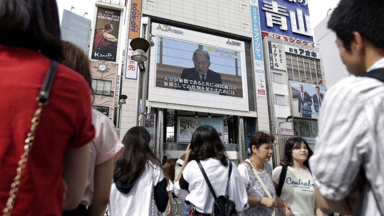 Kaiser Akihitos Ansprache wird auf einer Großbildleinwand in Tokio übertragen, davor stehen mehrere Menschen und hören zu. Kaiser Akihitos Ansprache wird auf einer Großbildleinwand in Tokio übertragen, davor stehen mehrere Menschen und hören zu.