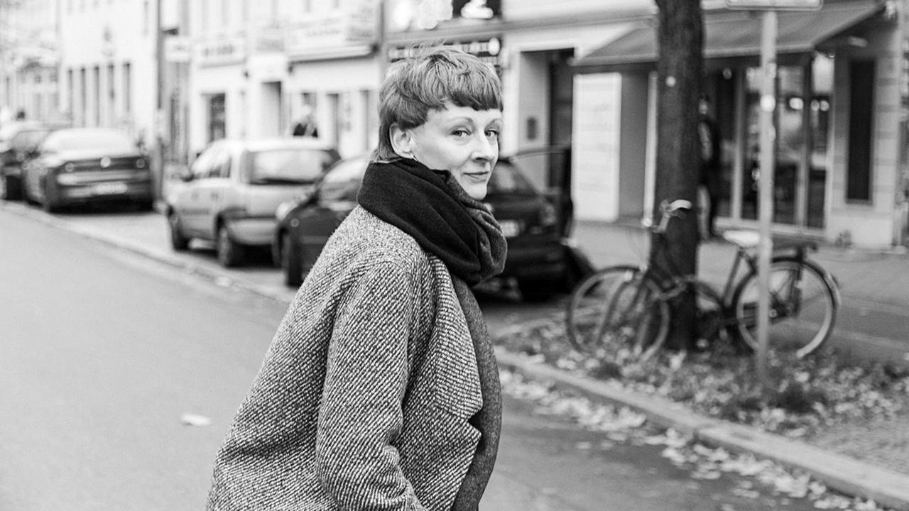 Alexandra Riedel ist auf einer Straße zu sehen, sie wendet ihren Kopf über die rechte Schulter halb zurück und blickt in Richtung Kamera. Sie trägt Mantel und hat einen Schal um, die Haare trägt sie kurz. Alexandra Riedel ist auf einer Straße zu sehen, sie wendet ihren Kopf über die rechte Schulter halb zurück und blickt in Richtung Kamera. Sie trägt Mantel und hat einen Schal um, die Haare trägt sie kurz.