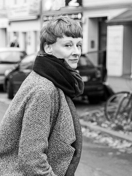 Alexandra Riedel ist auf einer Straße zu sehen, sie wendet ihren Kopf über die rechte Schulter halb zurück und blickt in Richtung Kamera. Sie trägt Mantel und hat einen Schal um, die Haare trägt sie kurz.