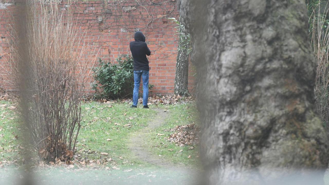 Ein Mann steht mit dem Rücken zur Kamera an einer Wand in einem Garten. Ein Mann steht mit dem Rücken zur Kamera an einer Wand in einem Garten.
