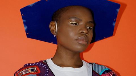 Pressebild der afroamerikanischen Künstlerin Vagabon vor orangenem Hintergrund. Sie trägt einen blauen Hut und farbenfrohe Kleidung.