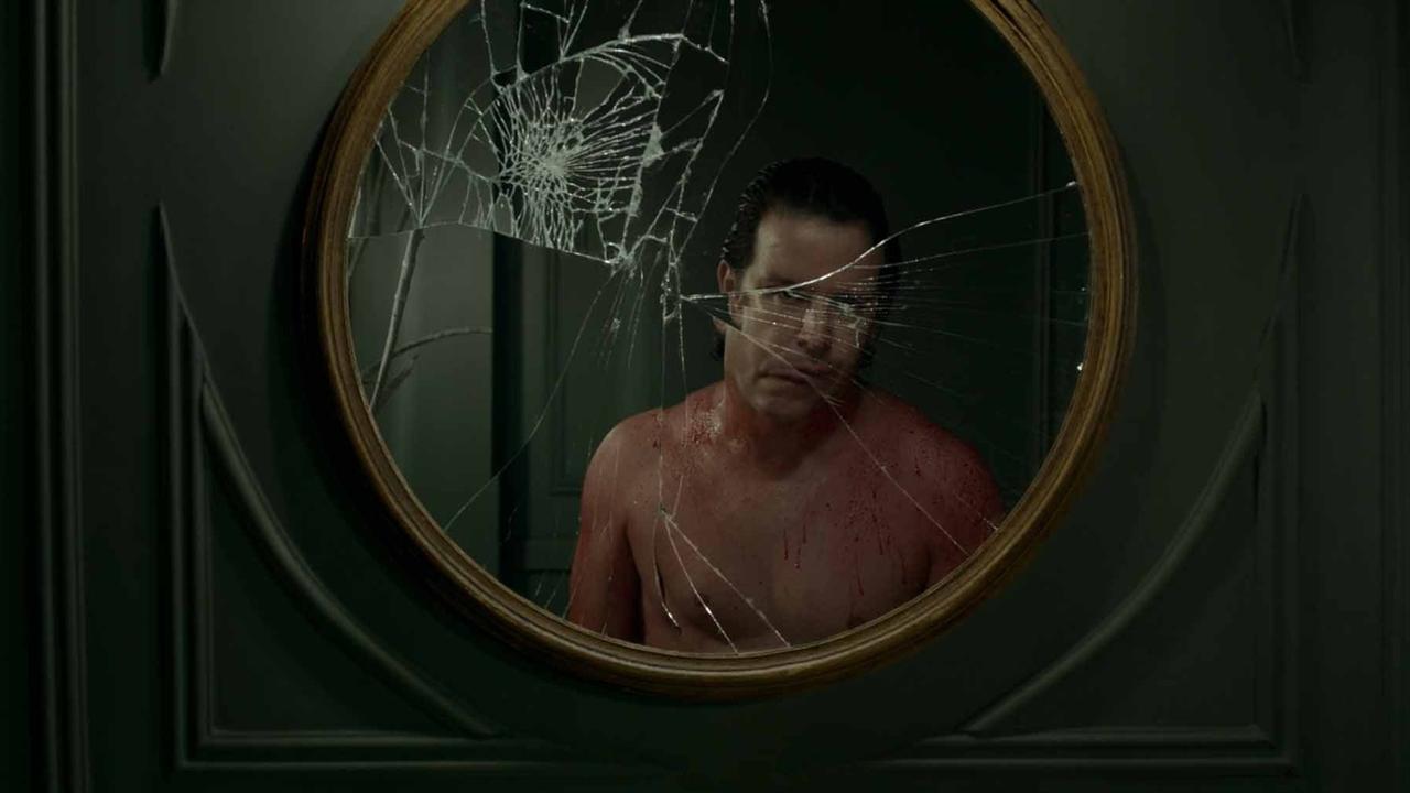 Ein Mann mit blutigem Oberkörper blickt in einen zersprungenen Spiegel.