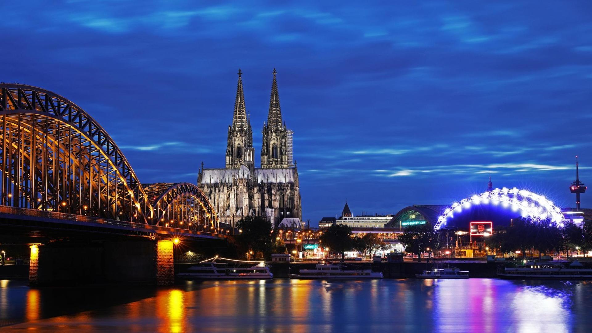 Das Foto zeigt den beleuchteten Dom in Köln neben der Hohenzollernbrücke. Das Foto zeigt den beleuchteten Dom in Köln neben der Hohenzollernbrücke.