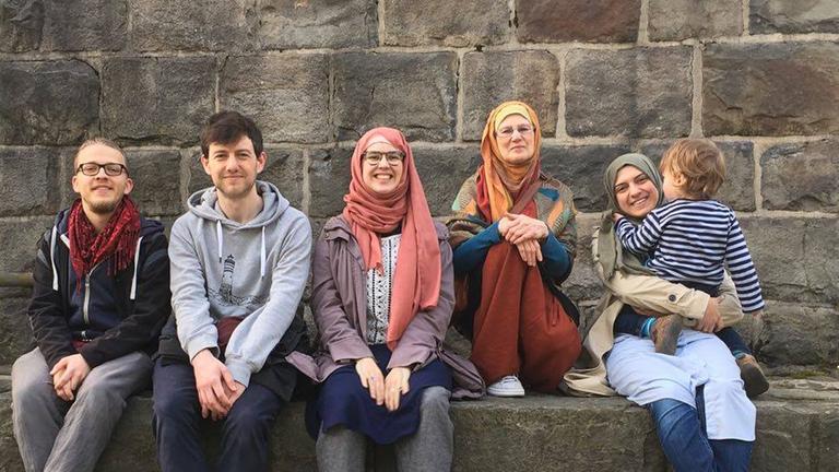 Zwei junge Männer und drei Frauen mit Kopftuch sitzen zusammen auf einer Mauer und schauen in die Kamera. Eine Frau hat ein Kleinkind im Arm. Zwei junge Männer und drei Frauen mit Kopftuch sitzen zusammen auf einer Mauer und schauen in die Kamera. Eine Frau hat ein Kleinkind im Arm.