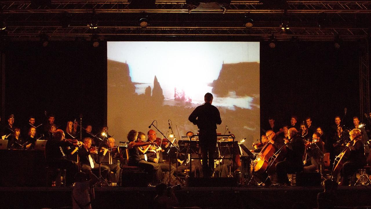 Ein Orchester mit Dirigent sind im Dunklen vor einer Leinwand zu sehen, auf der eine Brandung zu sehen ist. Links und rechts stehen im Hintergrund, neben der Leinwand, Sängerinnen und Sänger eines Chores. Ein Orchester mit Dirigent sind im Dunklen vor einer Leinwand zu sehen, auf der eine Brandung zu sehen ist. Links und rechts stehen im Hintergrund, neben der Leinwand, Sängerinnen und Sänger eines Chores.