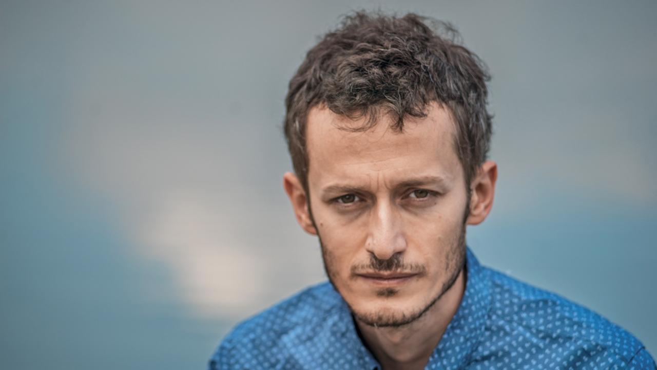 Der Autor Davit Gabunia schaut mit dem Gesicht in die Kamera. Er hat kurze, leicht gelockte dunkle Haare und trägt ein blaues Hemd. Der Hintergrund ist in türkis-blau gestaltet. Der Autor Davit Gabunia schaut mit dem Gesicht in die Kamera. Er hat kurze, leicht gelockte dunkle Haare und trägt ein blaues Hemd. Der Hintergrund ist in türkis-blau gestaltet.