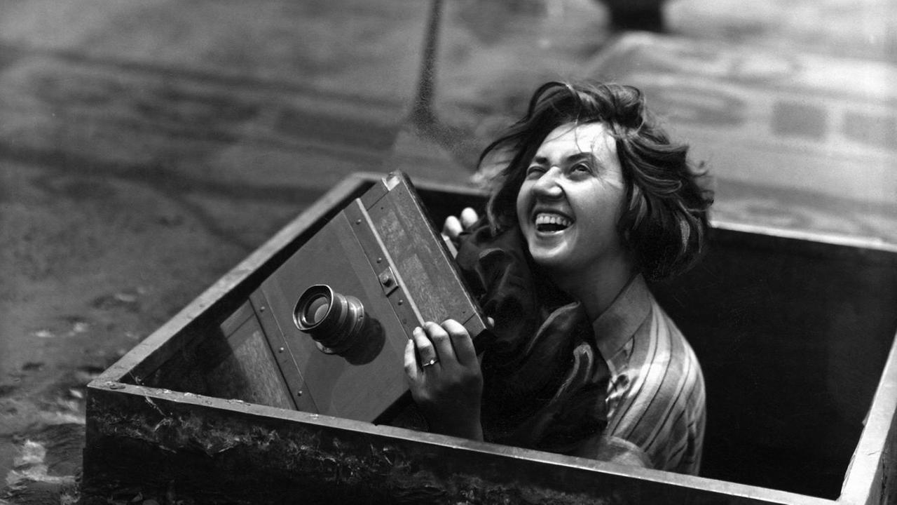 Die schwarz-weiß-Aufnahme einer jungen lachenden Frau, die mit einer alten Fotokamera aus einem Schacht herausschaut. Deutsches Reich: Aufnahme: B. Federmeyer - Erschienen in: 'Blatt' 26/1929 Originalaufnahme im Archiv von ullstein bild