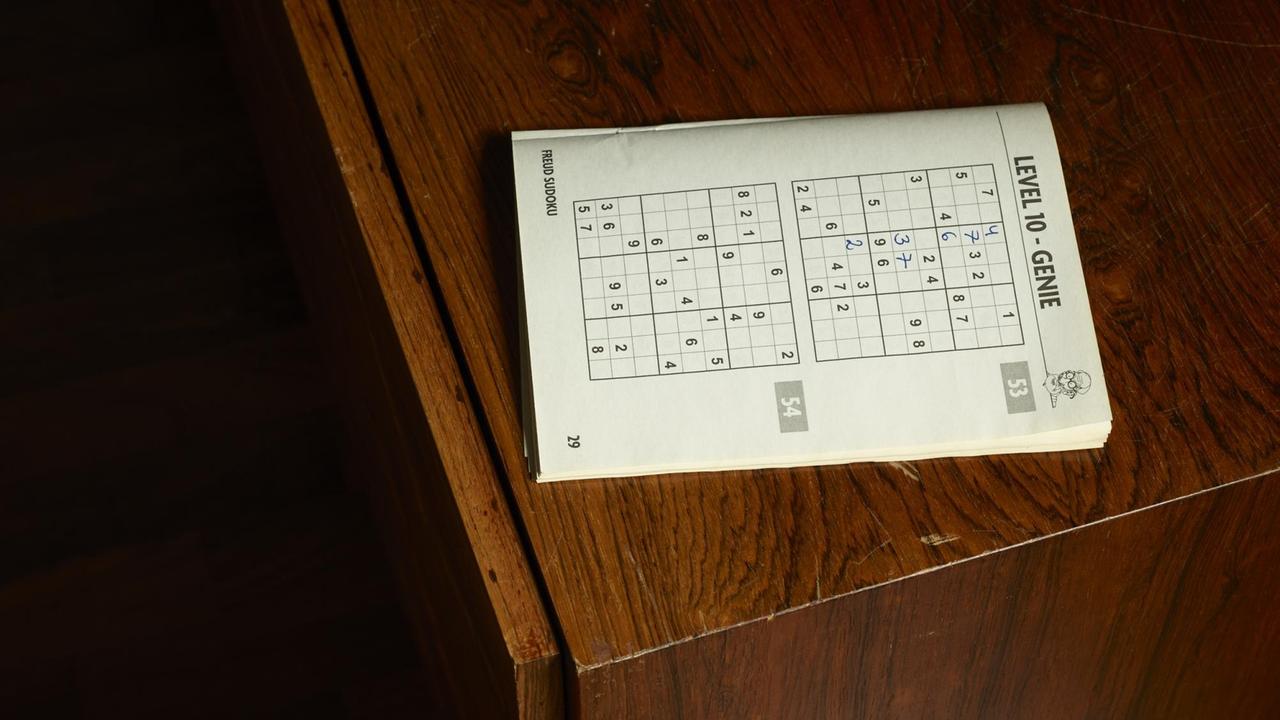 Ein aufgeschlagenes Rätselheft mit einem angefangenen Sudoku-Rätsel, Schwierigkeitsstufe "Genie", liegt auf einem dunkelbraunen Holzschrank.