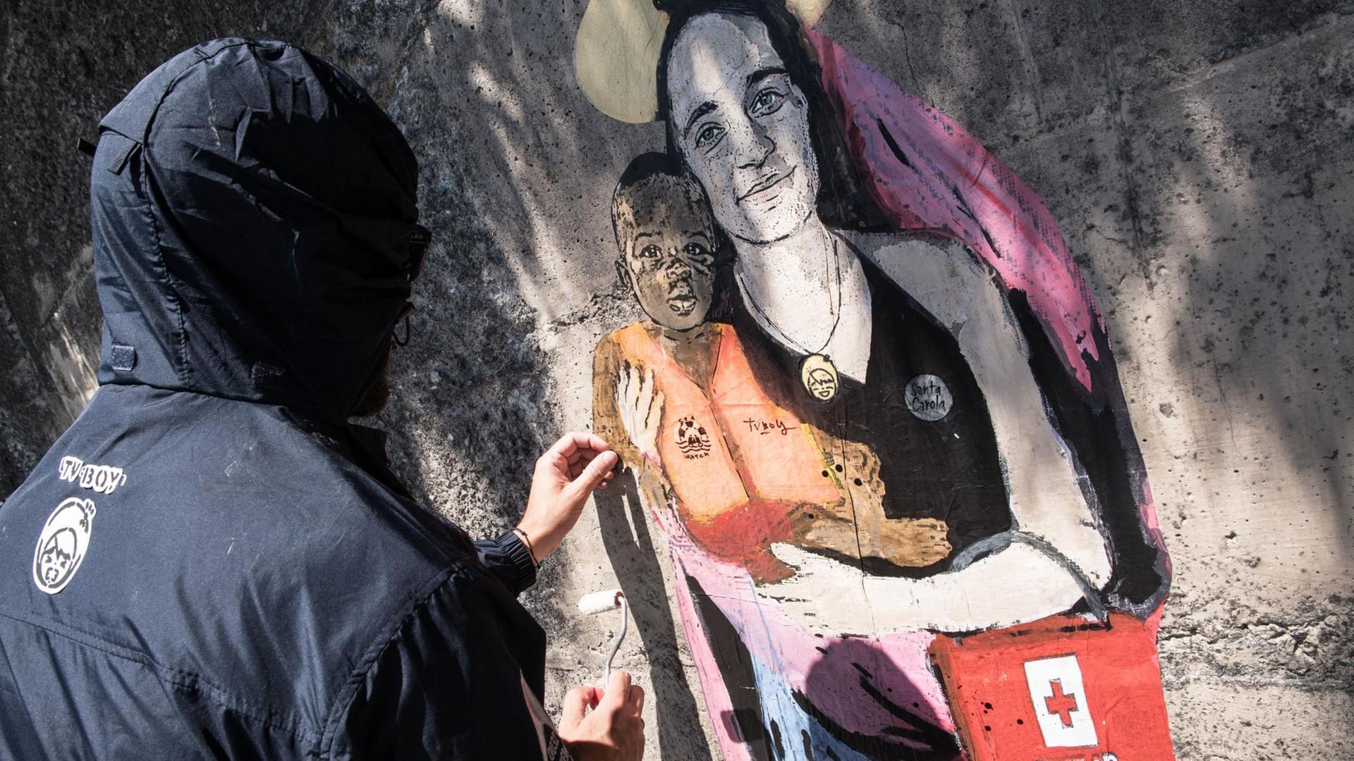 Der Street-Artist TVBOY malt sein Graffiti von Carola Rackete als Marienfigur an eine Wand in der sizilianischen Hafenstadt Taormina. Der Street-Artist TVBOY malt sein Graffiti von Carola Rackete als Marienfigur an eine Wand in der sizilianischen Hafenstadt Taormina.