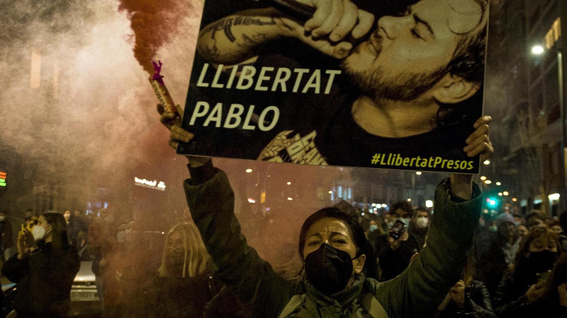 Demonstrierende in den Straßen von Barcelona fordern die Freilassung des spanischen Rappers Pablo Hasél, im Vordergrund hält eine Frau eine rote Rauchfackel und ein Plakat mit Porträt des Musikers und der Aufschrift "Llibertat Pablo" in die Höhe.