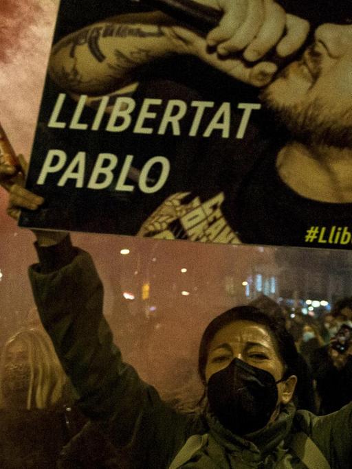Demonstrierende in den Straßen von Barcelona fordern die Freilassung des spanischen Rappers Pablo Hasél, im Vordergrund hält eine Frau eine rote Rauchfackel und ein Plakat mit Porträt des Musikers und der Aufschrift "Llibertat Pablo" in die Höhe.