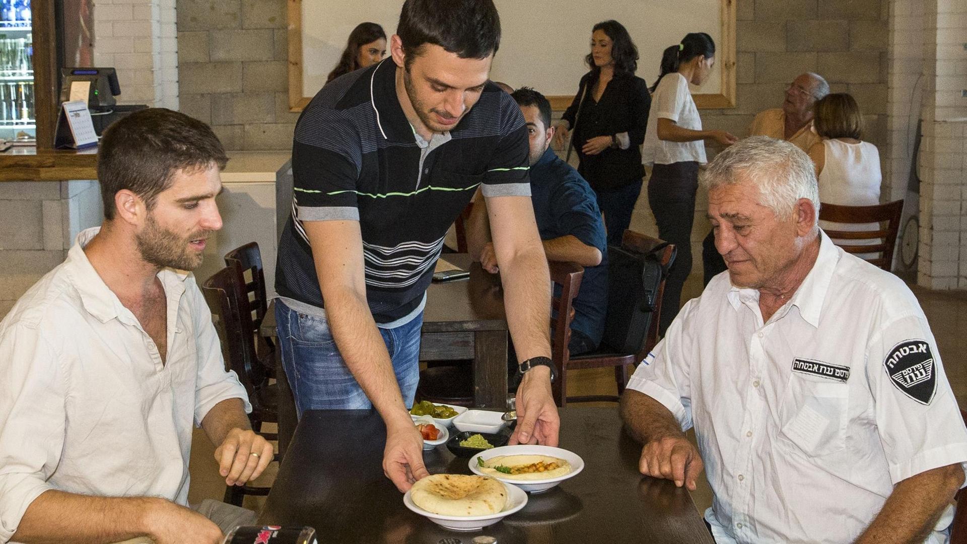 Der israelische Imbissbesitzer Kobi Tzafrir (m.) serviert seinen Gästen ein Gericht mit Kichererbsenpüree.