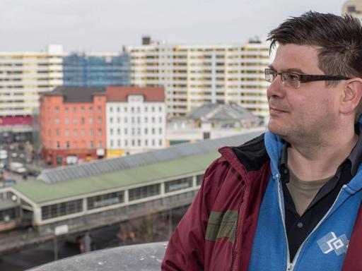 Andrej Holm steht auf einer Terrasse in Kreuzberg mit dem Wohnkomplex Zentrum Kreuzberg in seinem Rücken.