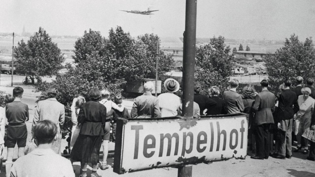 Passanten beobachten ein Flugzeug beim Landeanflug, im Vordergrund ein Schild mit der Aufschrift "Tempelhof". Passanten beobachten ein Flugzeug beim Landeanflug, im Vordergrund ein Schild mit der Aufschrift "Tempelhof".