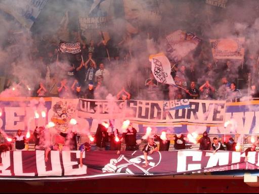 Bei einem Fußballspiel unterstützen Fans ihre Mannschaft auf den Rängen mit dem Abzünden von Bengalischen Feuern.