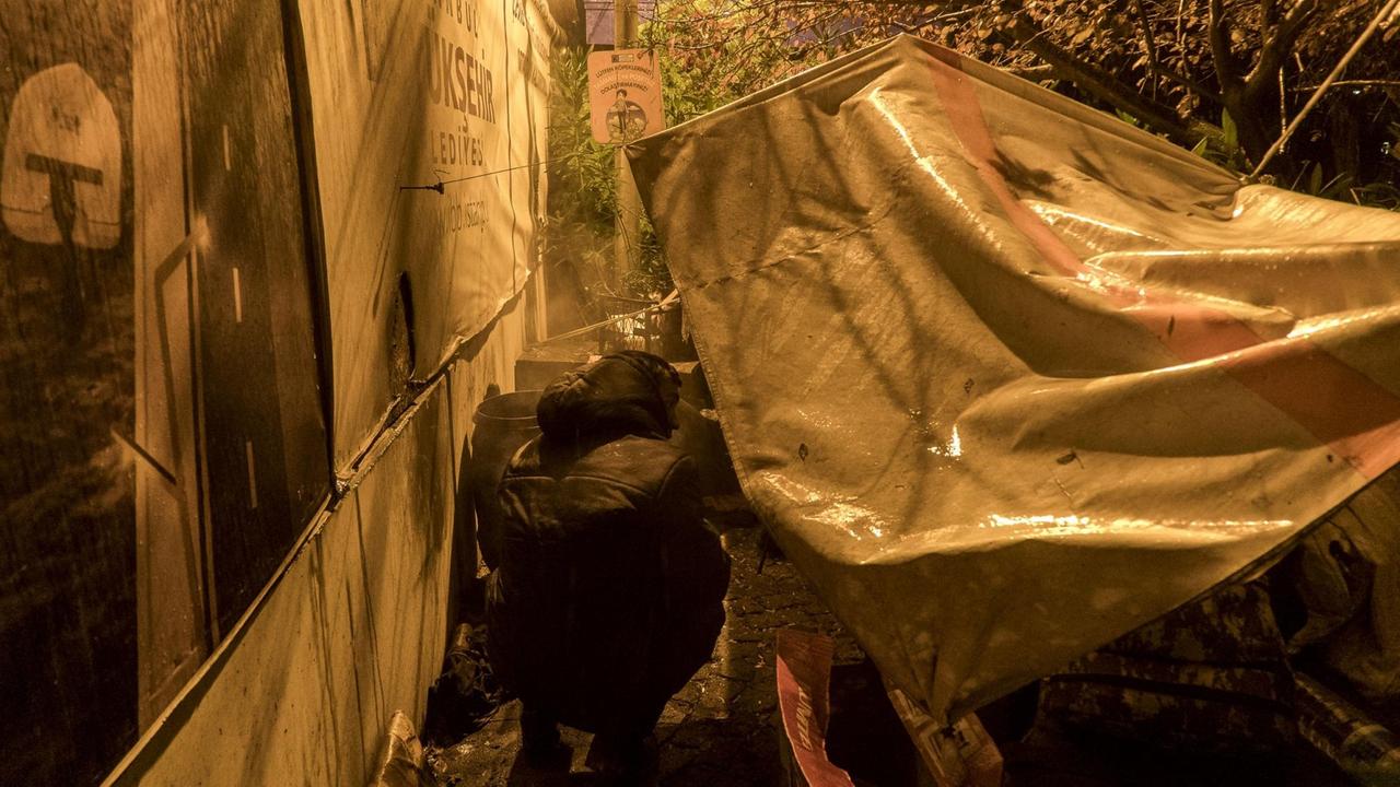 Ein Mann im Anorak, der nur von hinten zu sehen ist, kriecht unter eine große weiße regennasse Plane, die oben mit Seilen befestigt ist. Ein Mann im Anorak, der nur von hinten zu sehen ist, kriecht unter eine große weiße regennasse Plane, die oben mit Seilen befestigt ist.