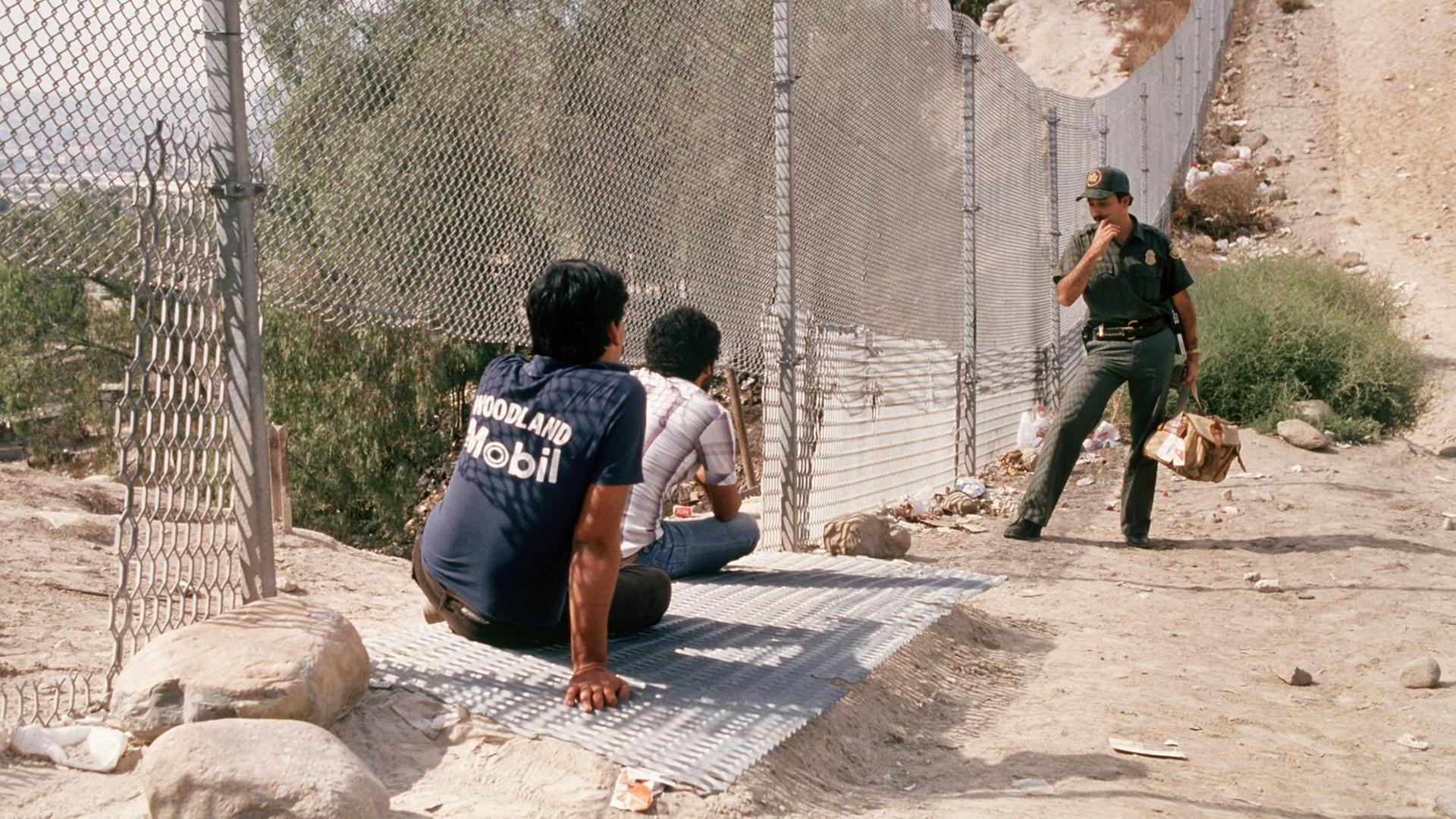 Ein US-Polizist steht an einem Grenzzaun. Vor ihm sitzen zwei Jugendliche im Sand.