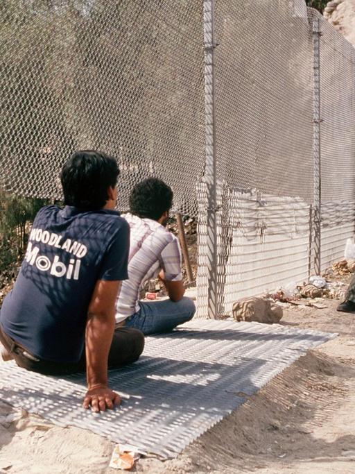 Ein US-Polizist steht an einem Grenzzaun. Vor ihm sitzen zwei Jugendliche im Sand.