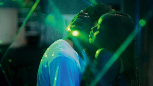Eine schwarze Frau und ein schwarzer Mann tanzen innig in einer dunkel-geheimnisvollen Atmosphäre, während grüne Lichtreflexe um sie herum blitzen.