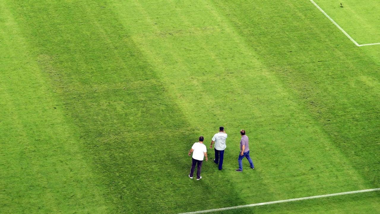 Drei Männer stehen auf dem Fußballfeld, auf dem nur bei genauem Hinschauen ein eingebranntes Hakenkreuz zu sehen ist
