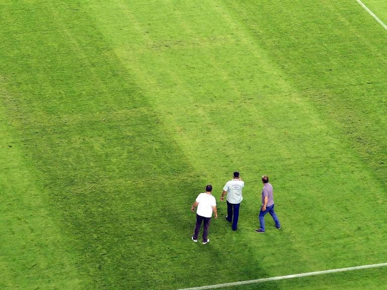 Drei Männer stehen auf dem Fußballfeld, auf dem nur bei genauem Hinschauen ein eingebranntes Hakenkreuz zu sehen ist
