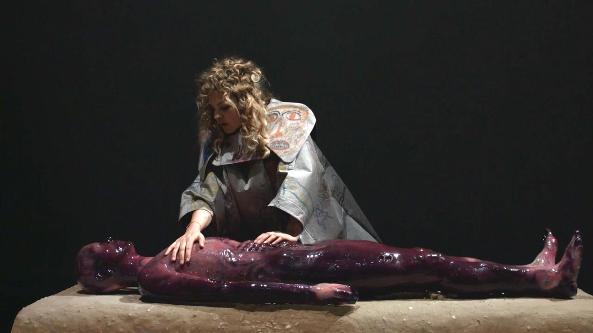 Eine Frau in einer Art Schamanengewand streicht über einen mit dunkelroter Flüssigkeit bedeckten Körper.