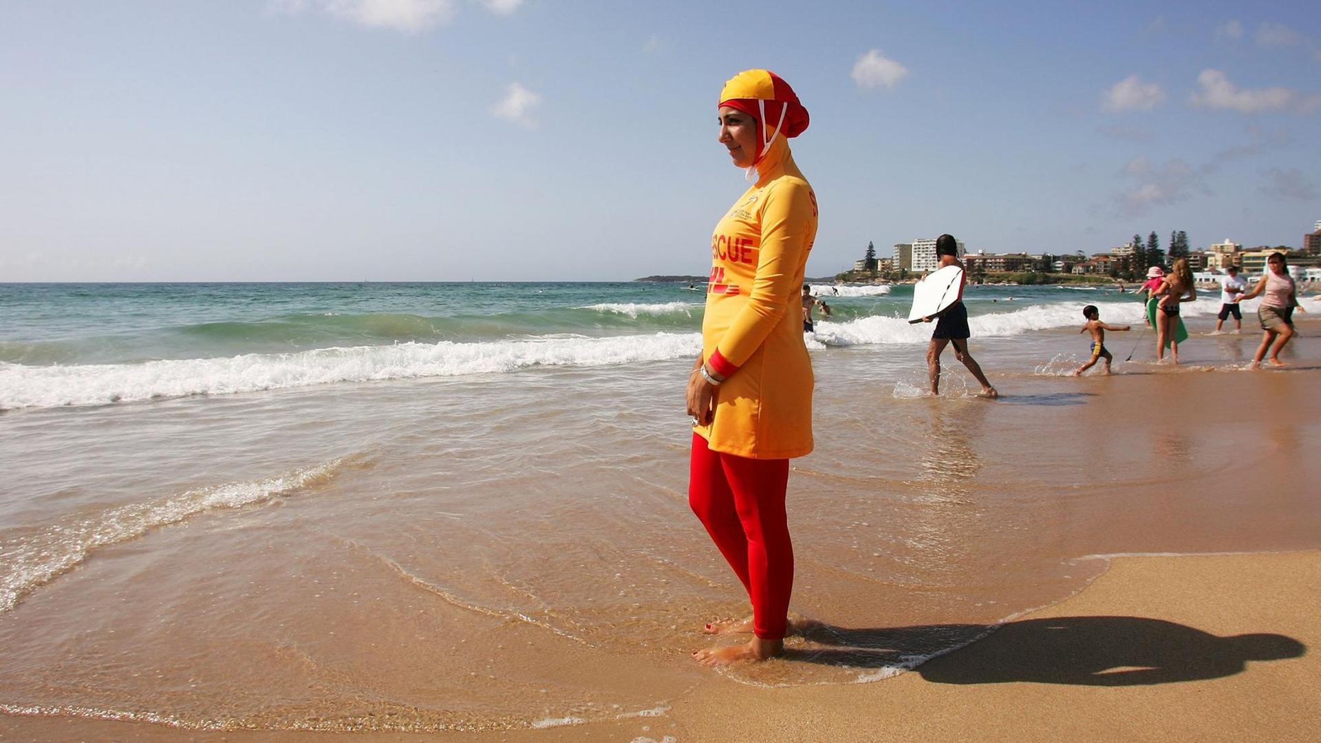 Eine muslimische Rettungsschwimmerin steht in einem Burkini am Strand von Sydney und schaut aufs Meer. Der Burkini ist rot und gelb. Im Hintergrund gehen Menschen mit Surfbrettern ins Wasser. Eine muslimische Rettungsschwimmerin steht in einem Burkini am Strand von Sydney und schaut aufs Meer. Der Burkini ist rot und gelb. Im Hintergrund gehen Menschen mit Surfbrettern ins Wasser.