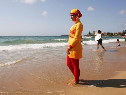 Eine muslimische Rettungsschwimmerin steht in einem Burkini am Strand von Sydney und schaut aufs Meer. Der Burkini ist rot und gelb. Im Hintergrund gehen Menschen mit Surfbrettern ins Wasser.