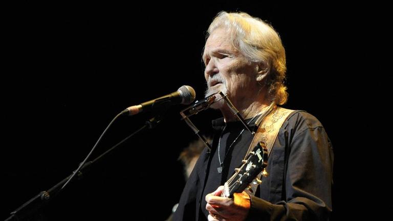 Kris Kristofferson steht auf der Bühne vor einem Mikrophon und spielt seine Gitarre, während er singt. Um seinen Hals hängt eine Mundharmonika-Halterung, die ihm einen schnellen Wechsel zum Spiel des Instrumentes ermöglicht. Kris Kristofferson steht auf der Bühne vor einem Mikrophon und spielt seine Gitarre, während er singt. Um seinen Hals hängt eine Mundharmonika-Halterung, die ihm einen schnellen Wechsel zum Spiel des Instrumentes ermöglicht.