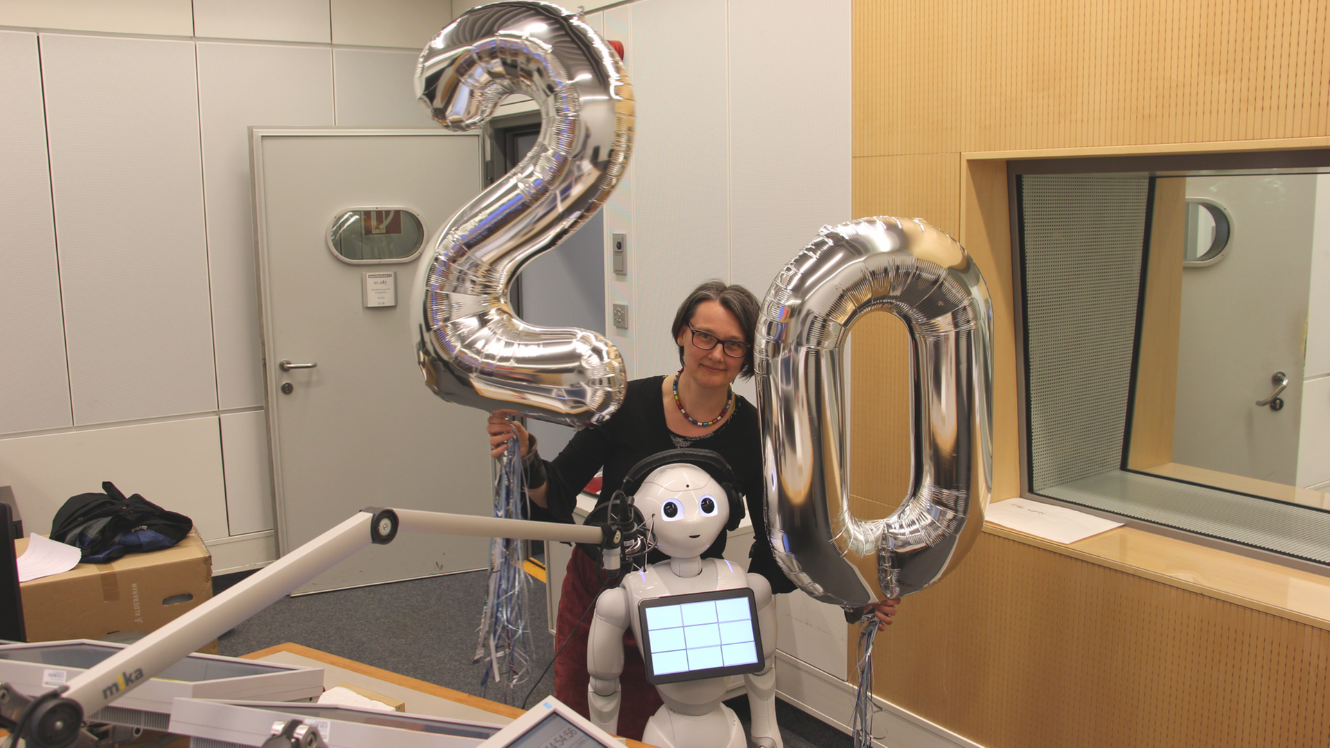 Eine Frau und ein kleiner weißer Roboter stehen im Sendestudio, die Frau hält zwei Ballons in Form einer "20" in den Händen. Eine Frau und ein kleiner weißer Roboter stehen im Sendestudio, die Frau hält zwei Ballons in Form einer "20" in den Händen.