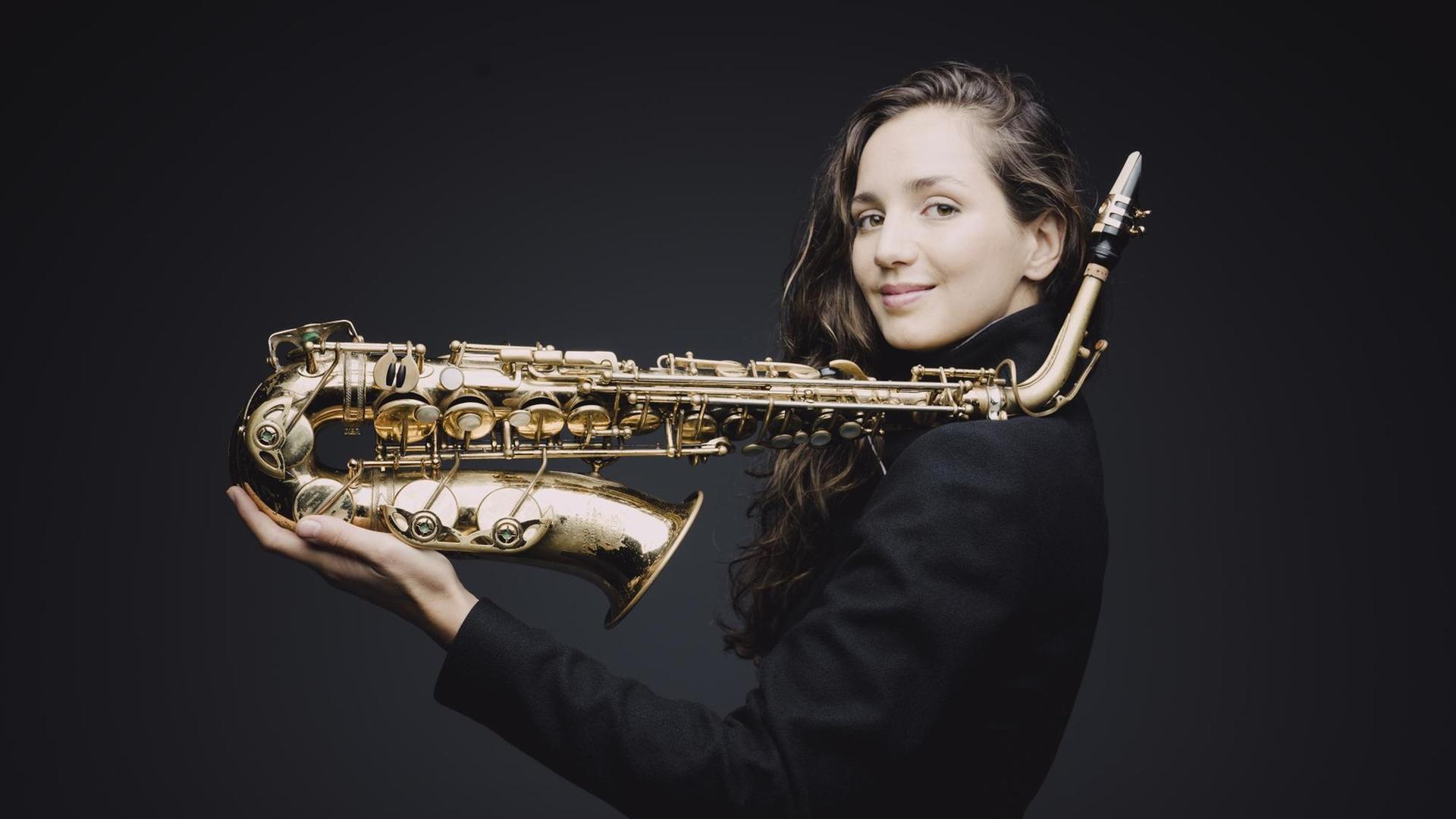 Eine junge Frau, mit langen braunen Haaren, steht seitlich vor einen schwarzen Hintergrund. In der Hand hält sie ihr Saxofon, der obere Teil des Instruments ist auf ihrer Schulter abgelegt. Eine junge Frau, mit langen braunen Haaren, steht seitlich vor einen schwarzen Hintergrund. In der Hand hält sie ihr Saxofon, der obere Teil des Instruments ist auf ihrer Schulter abgelegt.