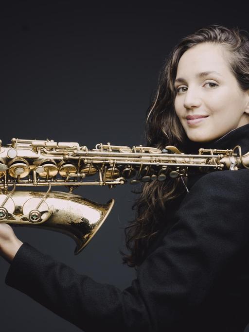 Eine junge Frau, mit langen braunen Haaren, steht seitlich vor einen schwarzen Hintergrund. In der Hand hält sie ihr Saxofon, der obere Teil des Instruments ist auf ihrer Schulter abgelegt.