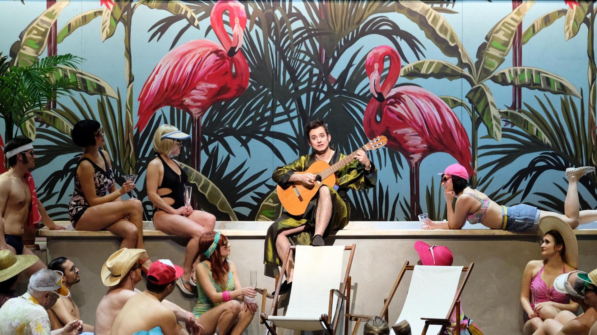 Ein Sänger sitzt mit Gitarre am Rand eines Swimmingpools, umgeben von Männern und Frauen in Badesachen. Hinter ihm sieht man zwei große gemalte Flamingos.