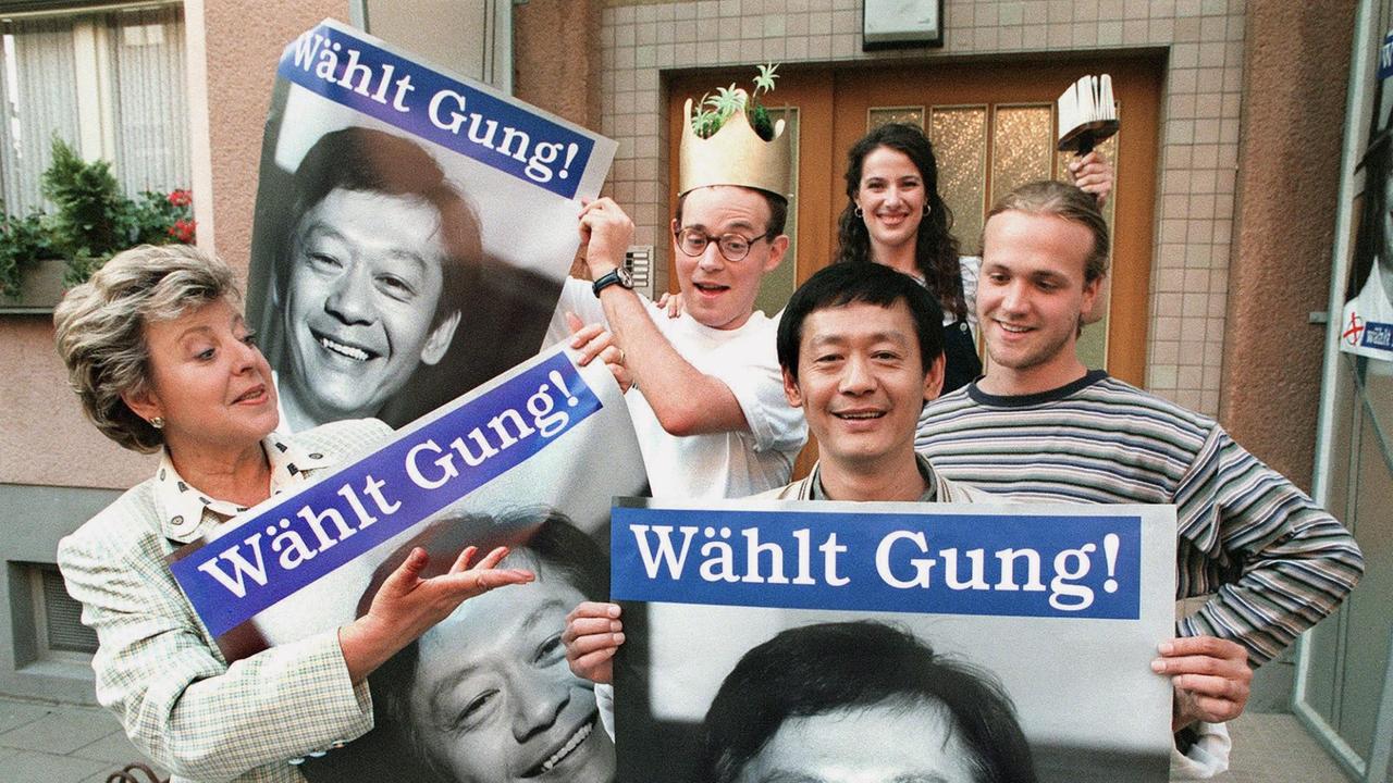 Szene aus Folge 667 der Lindenstraße: Die Schauspieler Marie-Luise Marjan, Philipp Neubauer, Amorn Surangkanjanajai, Clelia Sarto und Moritz A. Sachs halten Wahlplakate mit der Aufschrift "Wählt Gung" in ihren Händen. Szene aus Folge 667 der Lindenstraße: Die Schauspieler Marie-Luise Marjan, Philipp Neubauer, Amorn Surangkanjanajai, Clelia Sarto und Moritz A. Sachs halten Wahlplakate mit der Aufschrift "Wählt Gung" in ihren Händen.