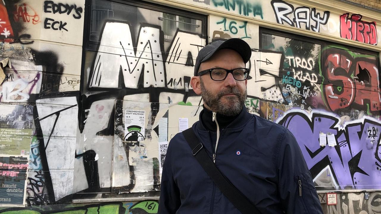 Der Connewitzer Sascha Lange mit Baseballkappe, Hornbrille und Bart vor einer mit Graffiti besprühten Hauswand in Leipzig-Connewitz.
