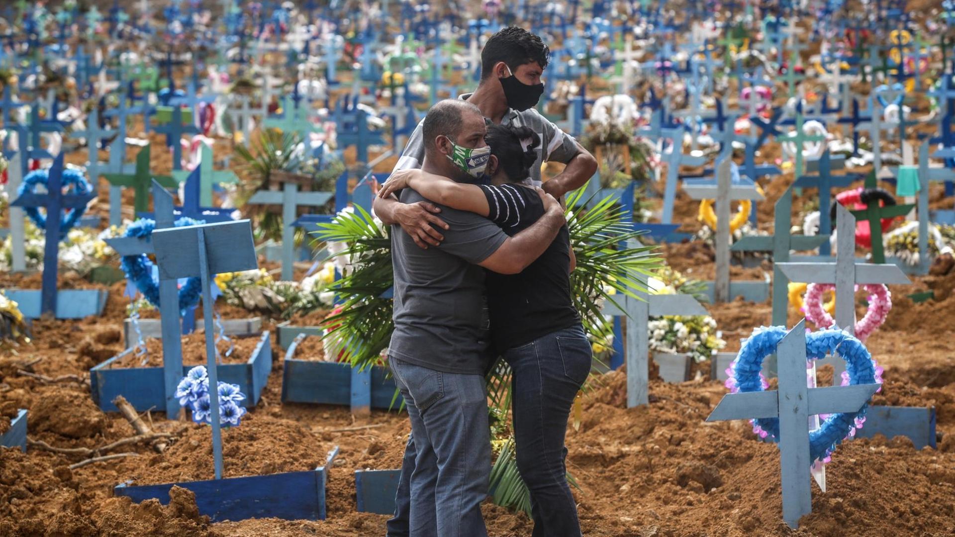 Drei Personen stehen dicht beieinander und umarmen sich, im Hintergrund sind viele Holzkreuze mit bunten Blumenkränzen zu sehen.