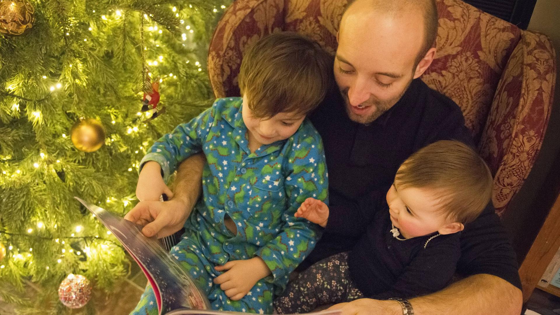 Ein Vater liest mit seinen zwei Kindern auf dem Schoß in einem Buch. Hinter ihnen steht der Weihnachtsbaum. Ein Vater liest mit seinen zwei Kindern auf dem Schoß in einem Buch. Hinter ihnen steht der Weihnachtsbaum.