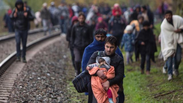 Viele Flüchtlinge laufen auf und entlang einer Bahnstrecke. Im Vordergrund trägt ein Mann ein in eine rote Decke eingewickeltes Kleinkind.
