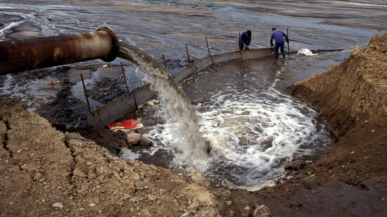 Abwasserrohr leitet Industrieabwässer in den verseuchten Bitterfelder Silbersee 1991, während 2 Männer im Wasser stehen und eine Barriere errichten. Abwasserrohr leitet Industrieabwässer in den verseuchten Bitterfelder Silbersee 1991, während 2 Männer im Wasser stehen und eine Barriere errichten.