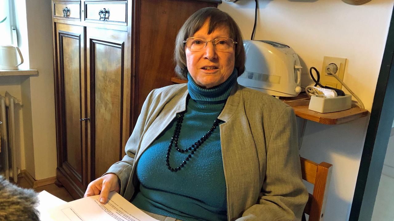 Porträtaufnahme von Ingrid Zasche. Eine ältere Dame mit kurzen, dunklen Haaren, randloser Brille. Sie trägt einen blauen Pullover, eine blaue Perlenkette und darüber ein grau-braunes Jackett. Porträtaufnahme von Ingrid Zasche. Eine ältere Dame mit kurzen, dunklen Haaren, randloser Brille. Sie trägt einen blauen Pullover, eine blaue Perlenkette und darüber ein grau-braunes Jackett.
