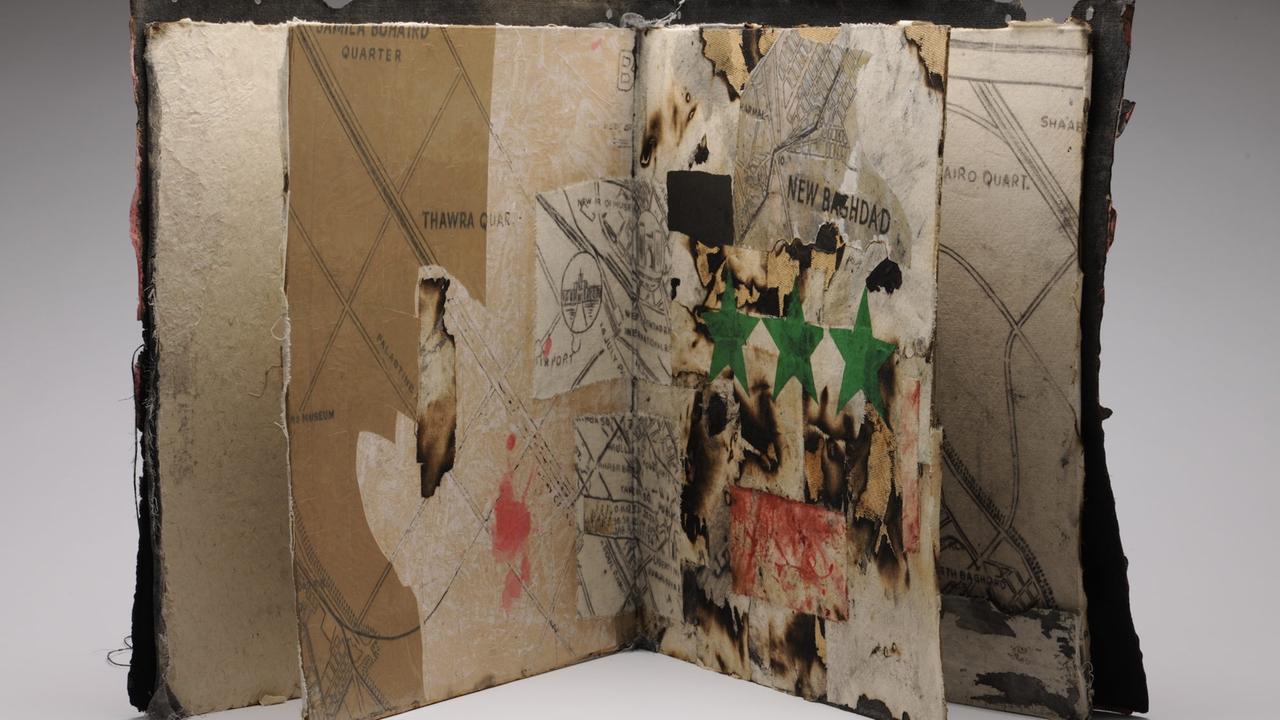 Ein aufgeschlagen aufgestelltes Notizbuch aus Holz, Tape und weiteren Materialien. Darin zu sehen sind Ausschnitte von Landkarten und grüne Sterne auf absichtlich verbrannten und geschwärzten Seiten. Ein aufgeschlagen aufgestelltes Notizbuch aus Holz, Tape und weiteren Materialien. Darin zu sehen sind Ausschnitte von Landkarten und grüne Sterne auf absichtlich verbrannten und geschwärzten Seiten.