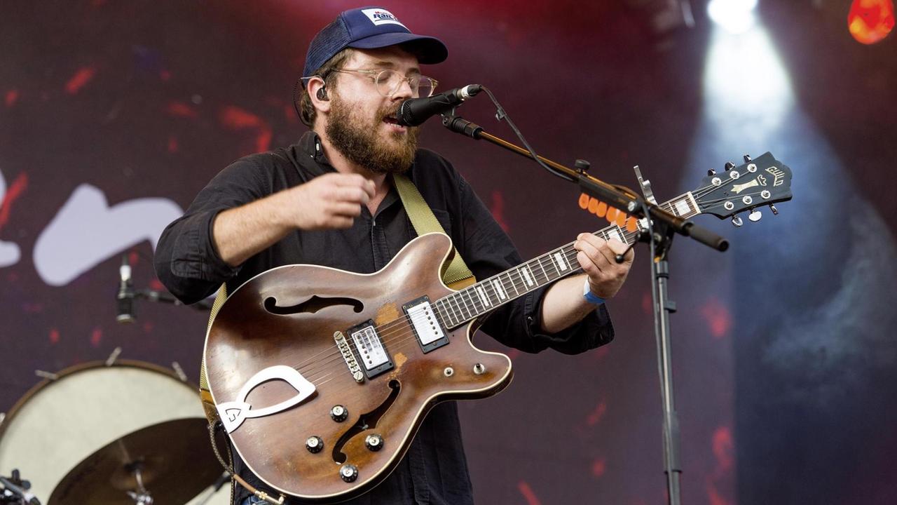 Mann mit blauer Kappe steht vor einem Mikrofon auf einer Bühne. Er hat seine Gitarre umgehangen. Mann mit blauer Kappe steht vor einem Mikrofon auf einer Bühne. Er hat seine Gitarre umgehangen.