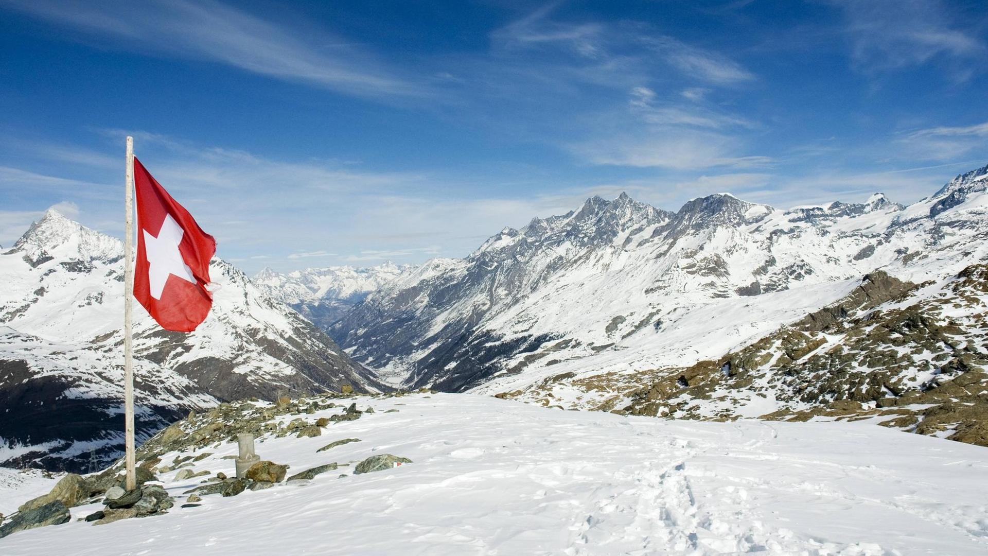 Eine Schweizer Fahne steckt im Schnee vor einem Alpenpanorama. Foto: imago stock&people Eine Schweizer Fahne steckt im Schnee vor einem Alpenpanorama. Foto: imago stock&people
