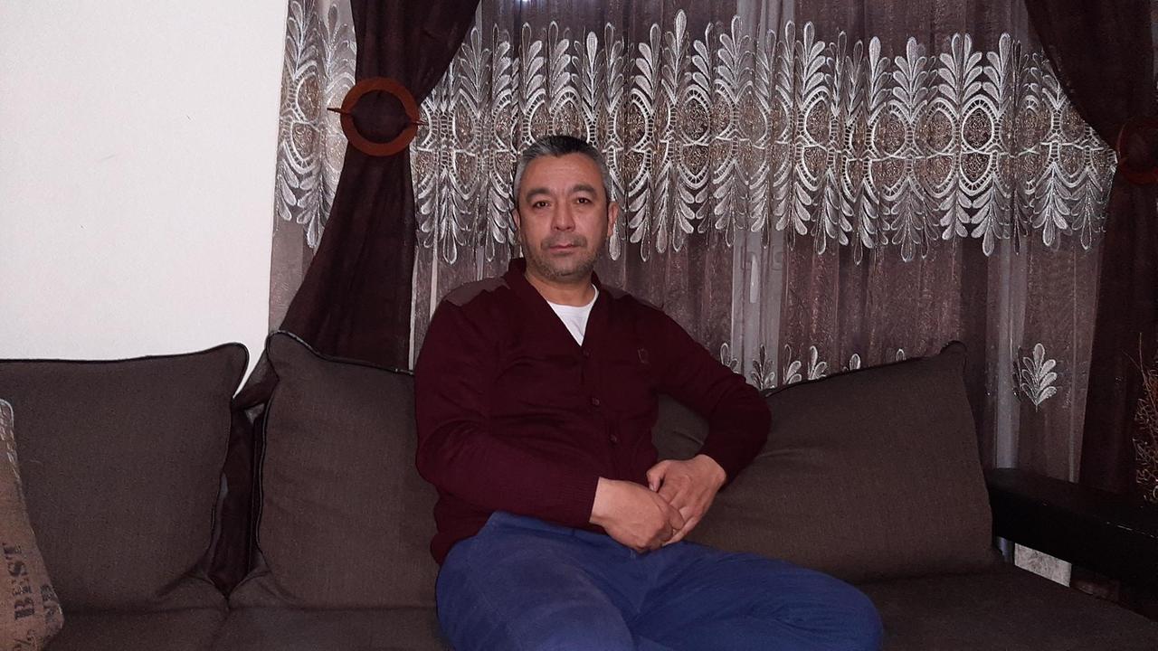 Ein Mann sitzt mit verschränkten Händen in Jeans und Pulli auf einem dunkelbraunen Sofa in einer Wohnung. Ein Mann sitzt mit verschränkten Händen in Jeans und Pulli auf einem dunkelbraunen Sofa in einer Wohnung.