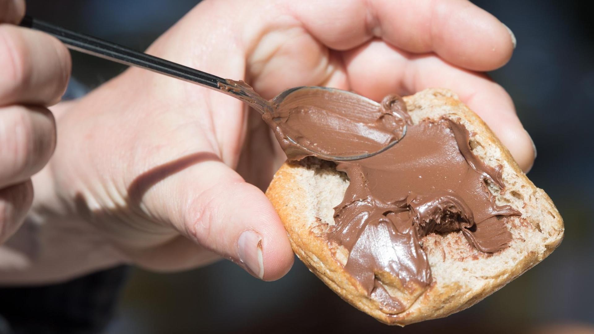 Eine Hand schmiert mit einem Löffel Nuss-Nougat-Creme auf ein Brötchen.