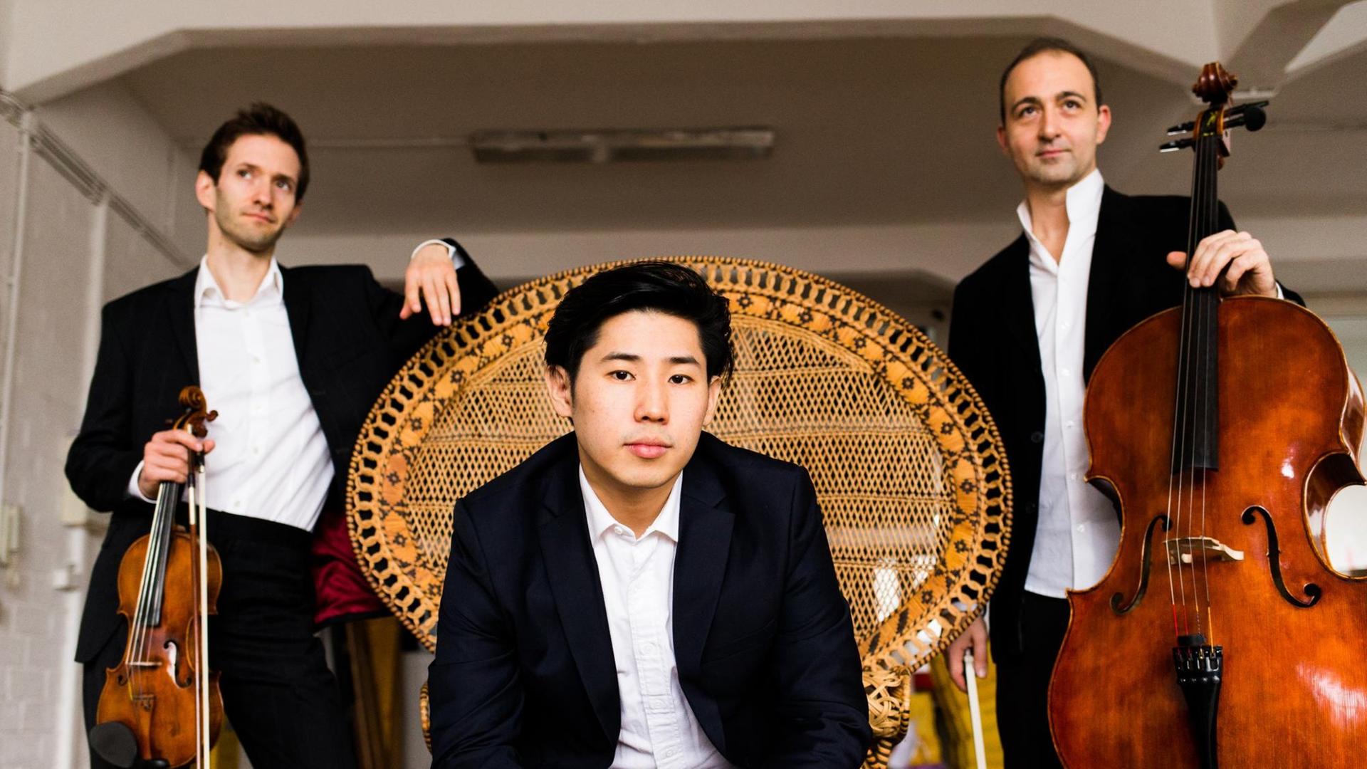 Pianist Jinsang Lee sitzt mittig auf einem großen Korbsessel, im Hintergrund stehen Geiger Mikhail Ovrutsky und Cellist Grigory Alumyan mit ihren Instrumenten in der Hand
