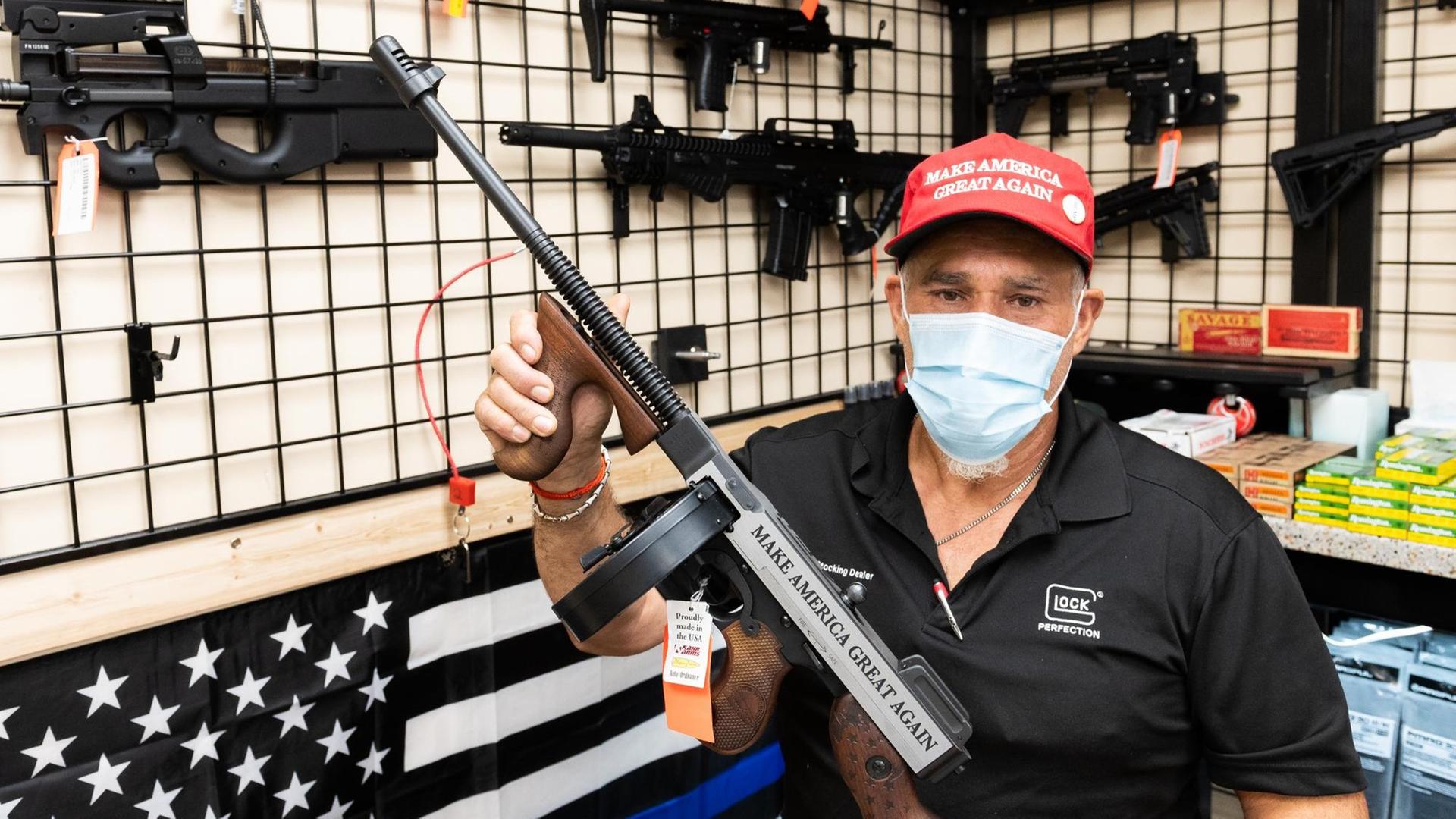 Ein Mitarbeiter eines Waffengeschäfts, mit Gesichtsmaske, präsentiert ein Gewehr. Auf der Waffe und auf der roten Basecap des Verkäufers ist der Schriftzug "Make America Great Again" zu sehen.