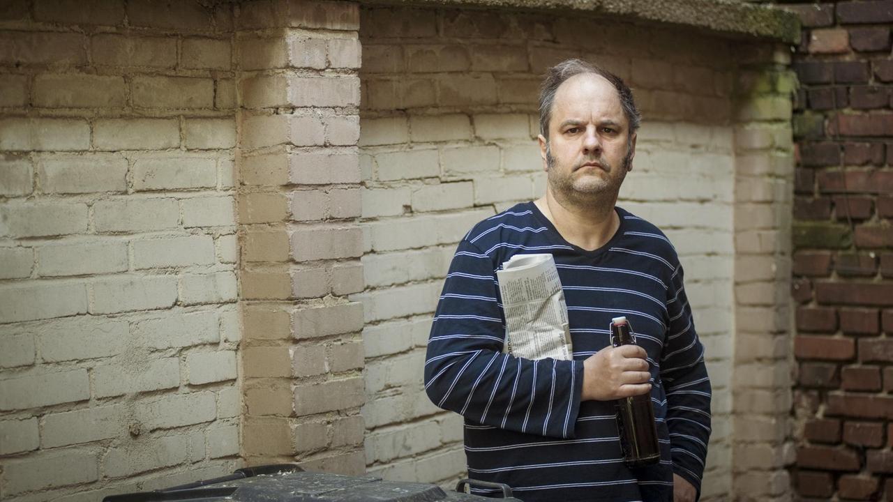 Der Kabarettist Matthias Egersdörfer steht im Schlafanzug mit einer Flasche Bier und einer Zeitung unterm Arm vor den Mülltonnen im Hinterhof eines Hauses und blickt ernst in die Kamera